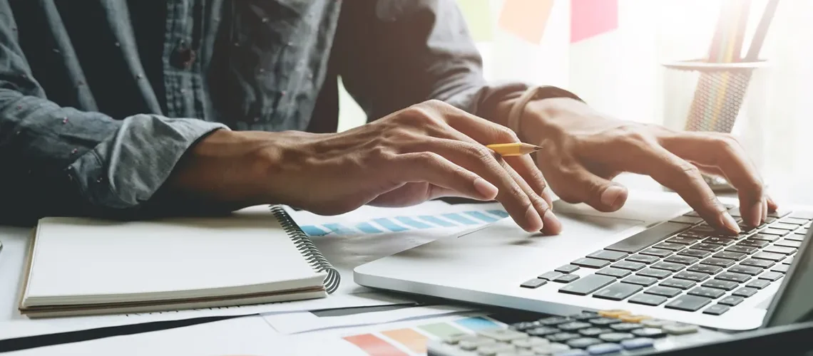 Close up of businessman or accountant hand holding pen working on laptop computer for calculate business data, accountancy document and calculator at office, business concept