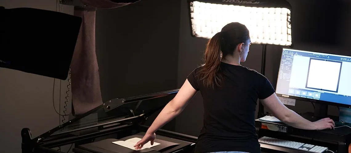 Technician Hannah Anderson reviews the digitized image of work she's scanning on a computer screen, holding her place in a book with her free hand.